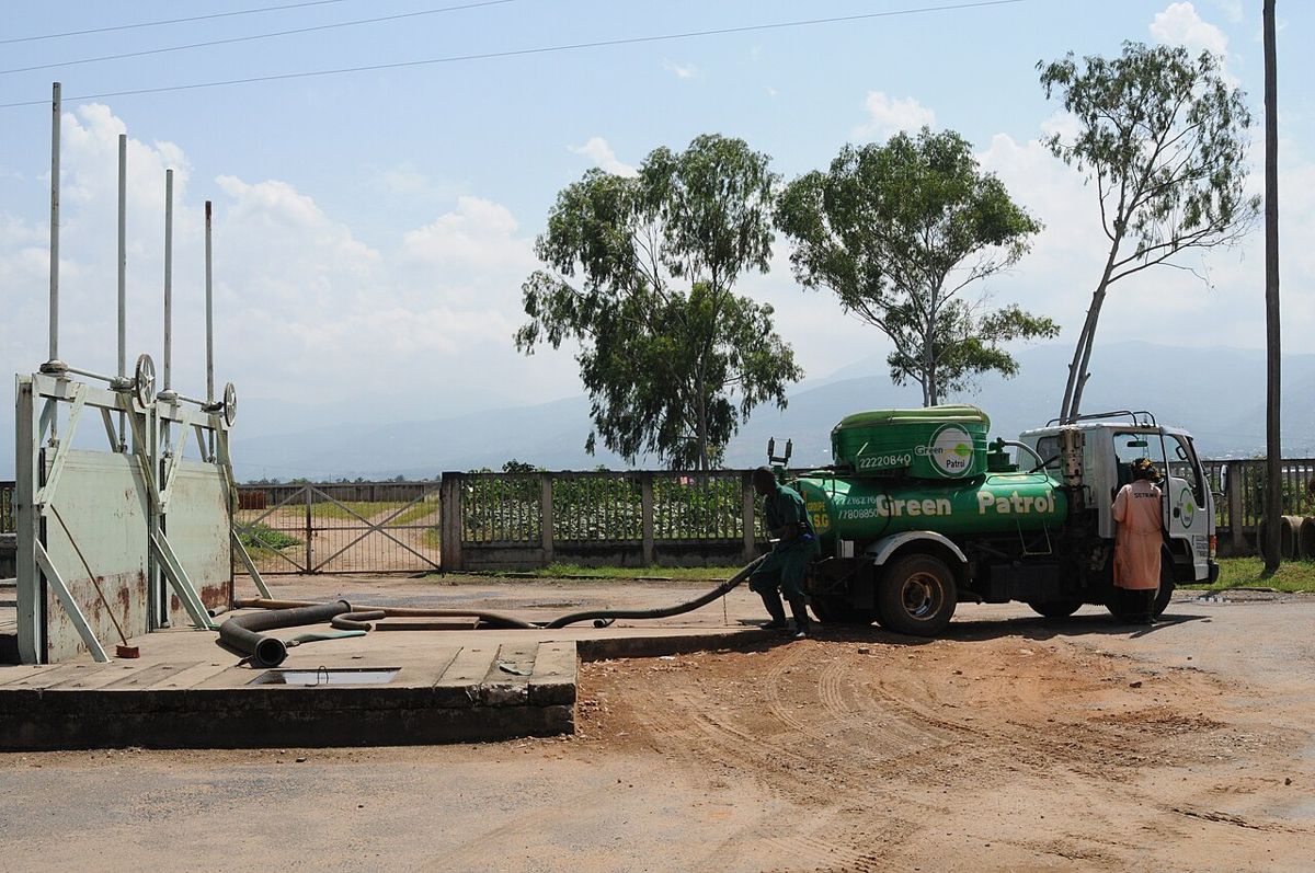 Truk vakum hijau dengan selang besar sedang mengosongkan muatan limbah septic tank, ditangani teknisi di lapangan terbuka